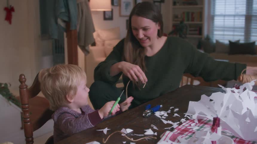 A young boy helps his mother cut twine with scissors as they happily make paper snowflake decorations for Christmas in slow motion