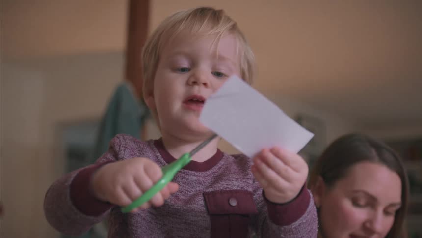 A young toddler carefully cuts a piece of paper with scissors in slow motion while his mother supervises