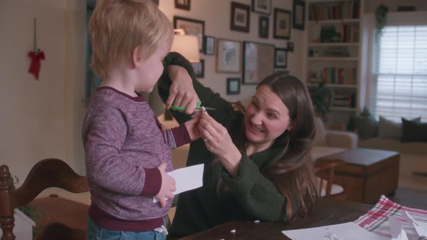 A mother and her young son make paper snowflakes together at a table in their cozy home during the Christmas season