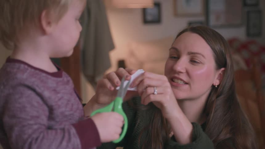 A smiling mother helps her excited toddler son learn to use scissors for the first time while making paper crafts at home