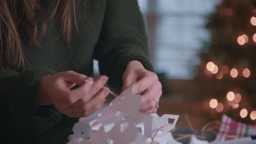 A mother and her child string together paper snowflakes for Christmas decorations in front of a twinkling tree