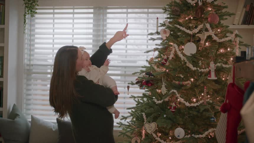 A mother holds her baby girl in her arms, pointing out the beautiful ornaments and lights on their Christmas tree