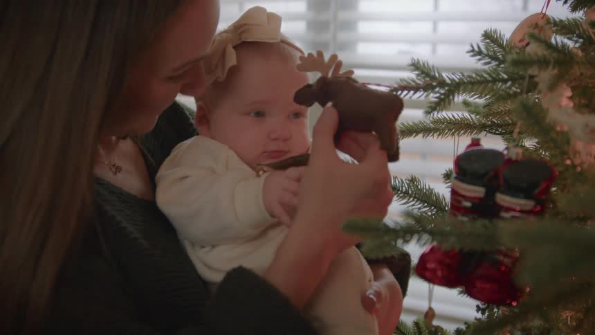 A mother holds her curious baby girl, showing her a moose ornament on the Christmas tree for the first time