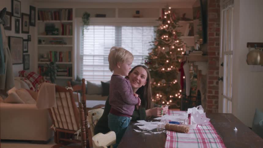A mother and her toddler son make paper snowflakes together at home during Christmas time, smiling and waving out the window