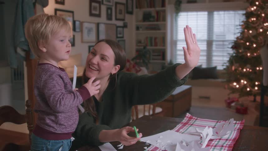 A joyful mother and her young son laugh and wave to family on a video call while making paper snowflakes for Christmas