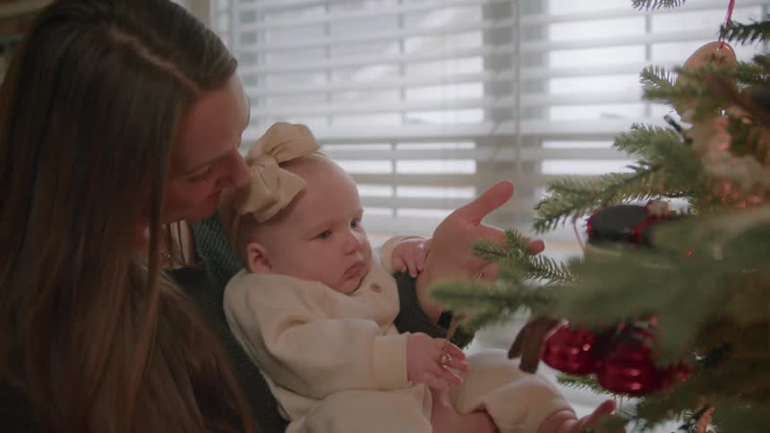 A loving mother holds her baby daughter, showing her the family Christmas tree and its ornaments for the first time