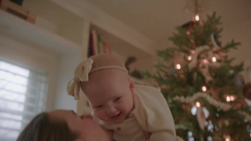 A mother playfully lifts her smiling baby girl and gives her a kiss in front of a festive Christmas tree