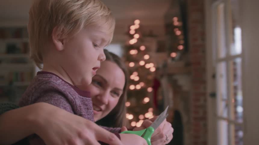 A mother helps her toddler son learn to use scissors for the first time in slow motion, with a Christmas tree in the background
