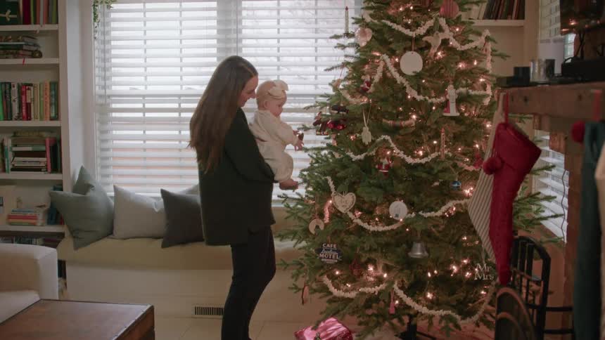 A mother holds her baby girl in her arms, letting her touch the ornaments on their festive Christmas tree