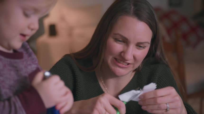 A smiling mother carefully cuts a paper snowflake with scissors as her young son watches in their cozy home