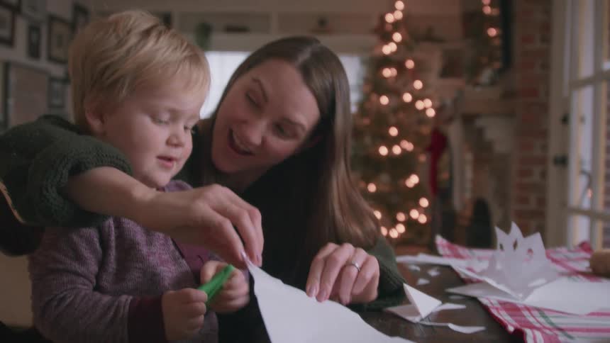A young mother lovingly helps her little boy as he learns to use scissors to cut out paper snowflakes for Christmas