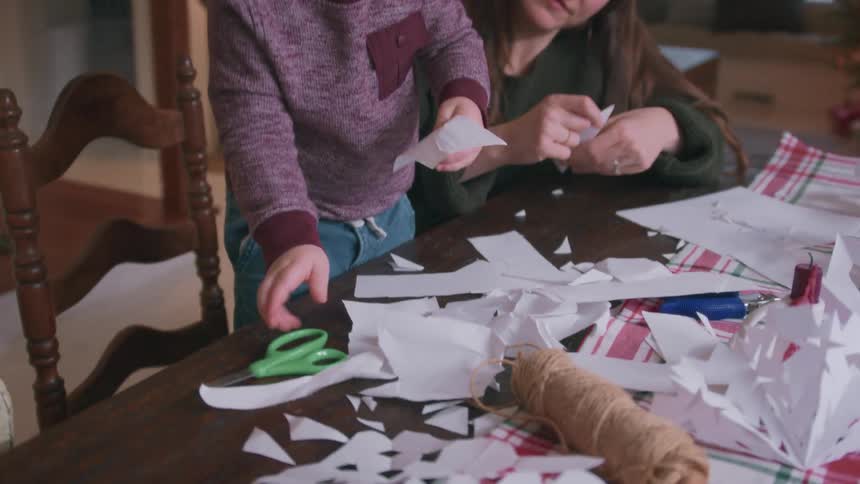 A mother and her young toddler son spend quality time together making paper snowflakes for Christmas at a messy craft table in their cozy home