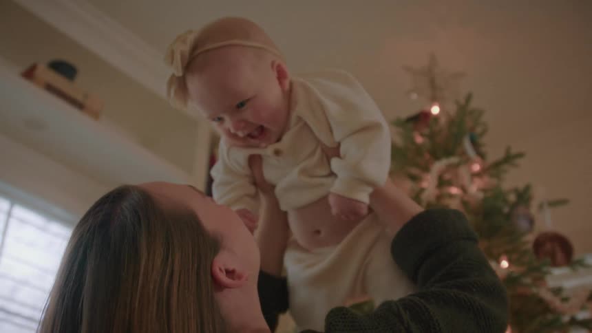 A mother joyfully lifts her laughing baby girl into the air in front of a lit Christmas tree