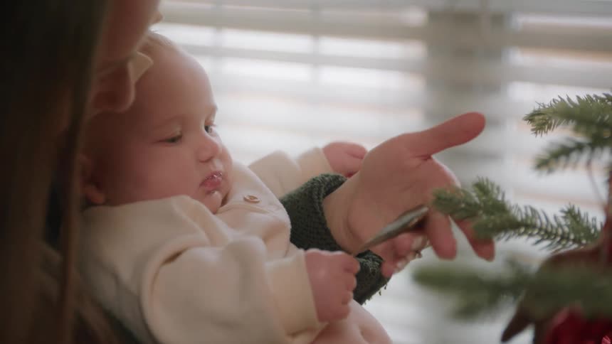 A mother gently holds her infant daughter as the baby curiously touches a heart-shaped ornament on a Christmas tree for the first time
