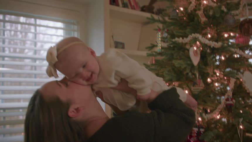 A mother lovingly lifts her joyful baby girl into the air in front of a twinkling Christmas tree in slow motion