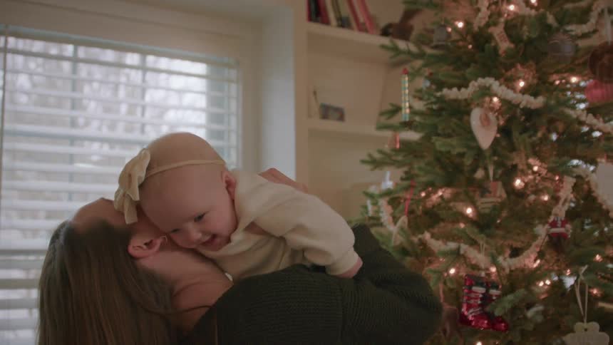 A loving mother playfully lifts her laughing baby girl in front of a lit Christmas tree before embracing her