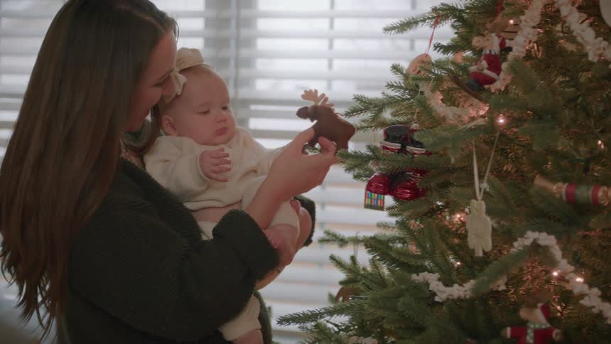 A loving mother holds her baby girl and shows her a cute moose ornament on their beautifully decorated Christmas tree
