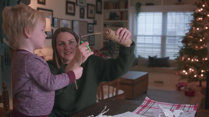A mother and her toddler son enjoy a festive afternoon crafting paper snowflakes and cutting twine for Christmas decorations in their cozy living room