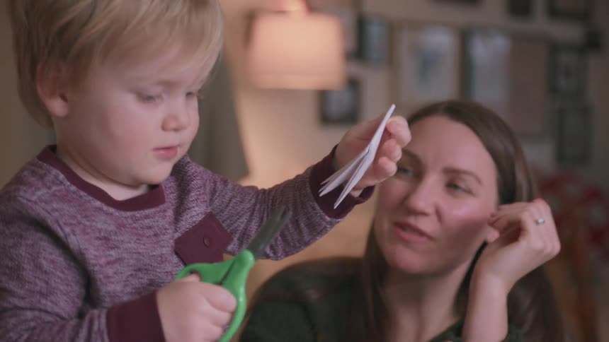 A mother patiently teaches her young blonde son how to use scissors while making paper crafts together at home