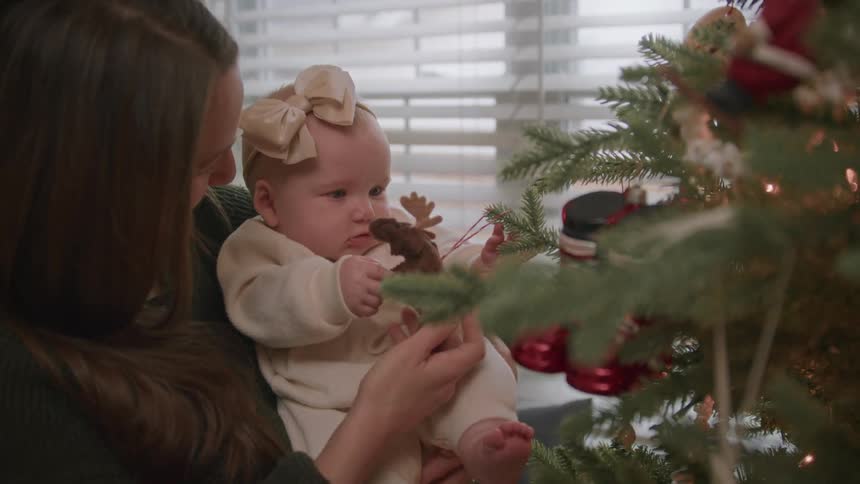 A mother holds her baby girl and shows her a moose ornament on their decorated Christmas tree