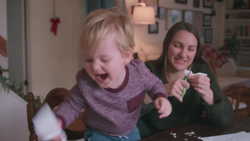 A joyful toddler laughs and plays with paper scraps while his mom cuts out paper snowflakes for Christmas decorations in their cozy home