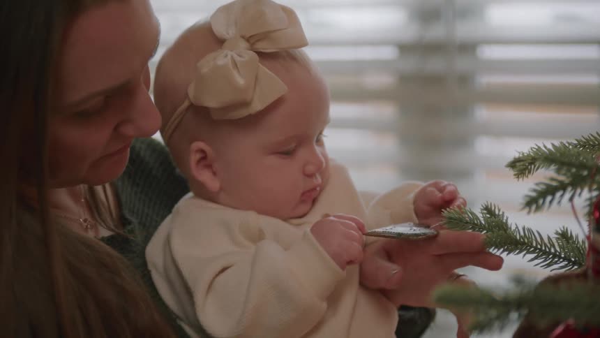 In slow motion, a mother holds her baby girl as she curiously explores the branches and a heart-shaped ornament on a Christmas tree