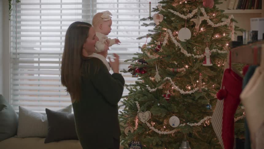 A loving mother holds her baby daughter, showing her the beautiful, lit-up Christmas tree for the first time