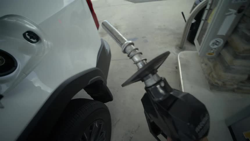 A first-person point-of-view of selecting premium unleaded gasoline and inserting the nozzle to refuel a white car in slow motion