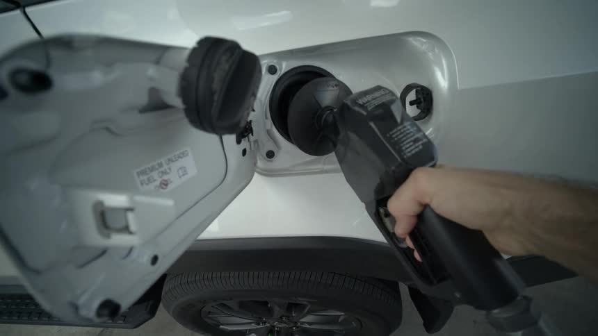 A first-person point-of-view shot of a hand taking a gas nozzle and pumping fuel into a white car at a gas station