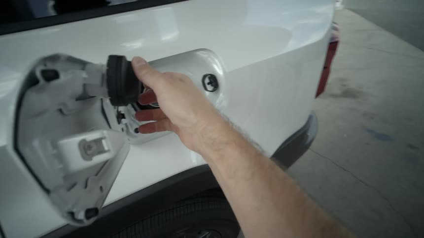 First-person perspective of pumping gas into a white vehicle at a gas station, returning the nozzle, and securing the fuel cap