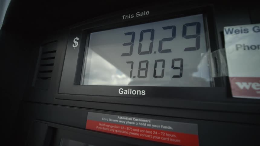 A close-up shot of a digital gas pump meter shows the rising cost and gallons of fuel being pumped into a vehicle