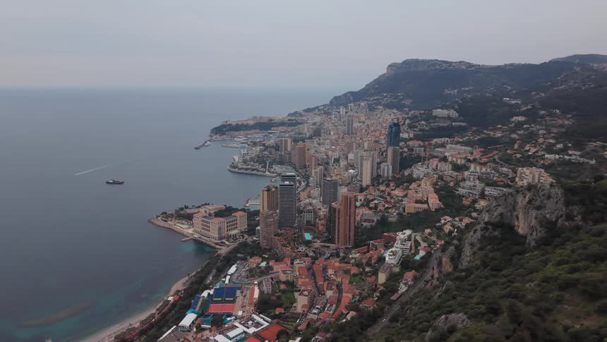 A wide aerial shot reveals the Principality of Monaco and its harbor nestled between the mountains and the sea on an overcast day