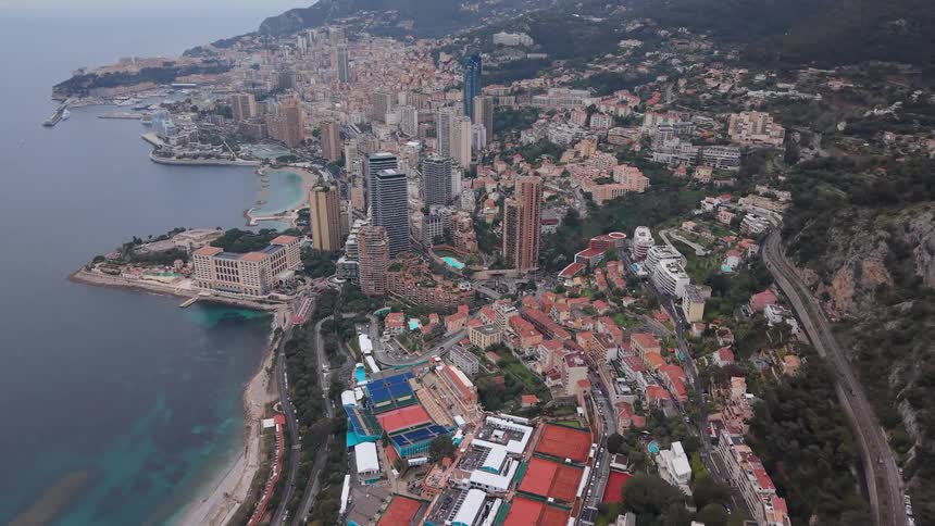 Expansive aerial view slowly pulls back over the Monte Carlo Country Club tennis courts to reveal the stunning cityscape of Monaco nestled between the mountains and the sea