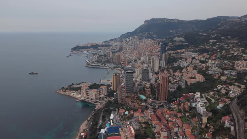 A static aerial view shows the cityscape of Monaco nestled between the mountains and the Mediterranean Sea on an overcast day