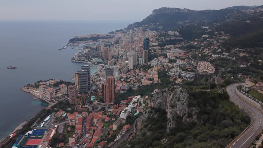 An expansive aerial view reveals the city of Monaco nestled between the mountains and the Mediterranean Sea on an overcast day