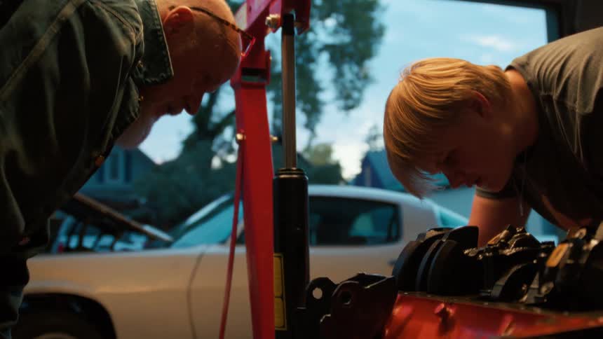 Side view of a grandfather pointing and instructing his grandson as they work on a car engine in the garage at dusk