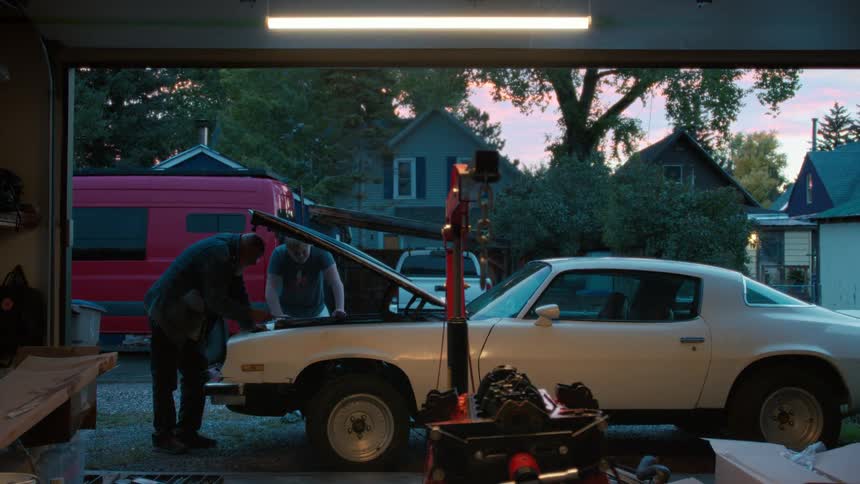 An older man and a younger man work together on the engine of a classic white sports car in their driveway at dusk