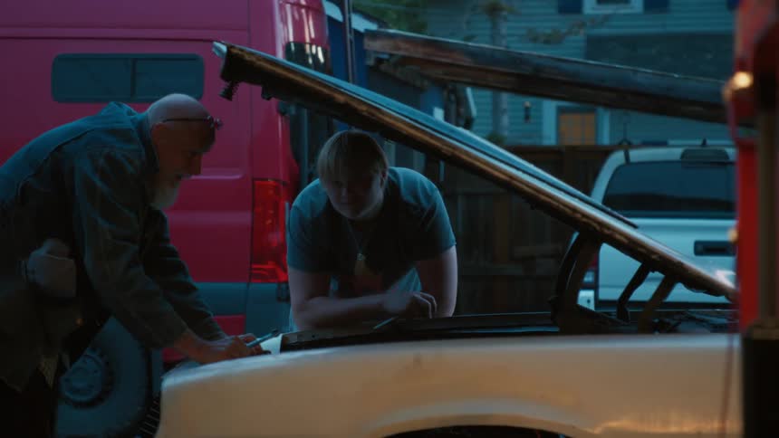 A senior mechanic with a white beard teaches a young man how to fix a car engine in their driveway at twilight