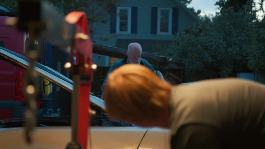 An older man with a white beard supervises a younger man as they work on a car engine together in a driveway at dusk