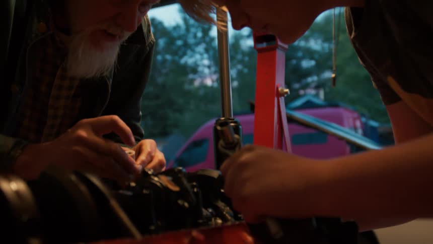 An elderly man with a white beard teaches a young person how to assemble a car engine in a home garage