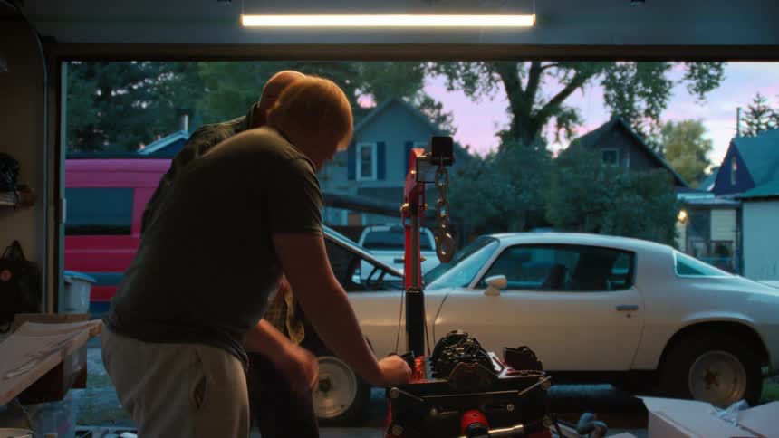 An older man and a younger man work together on a classic car engine in their home garage at dusk
