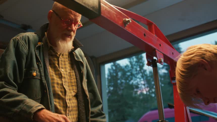 A seasoned mechanic with a white beard mentors a young apprentice as they work together on a car engine in a dimly lit garage