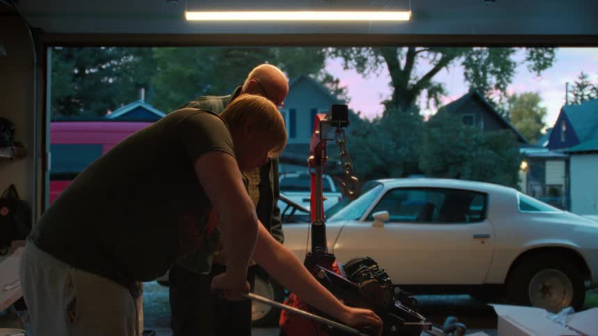 A grandfather teaches his grandson how to work on a classic car engine in their home garage at sunset