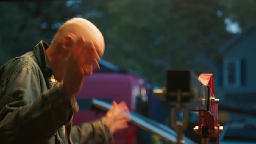 A frustrated senior mechanic gestures in slow motion while working on a car with an engine hoist in his garage at dusk