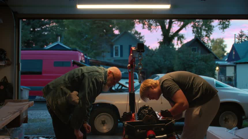A grandfather teaches his grandson how to work on a classic car engine in their home garage at twilight