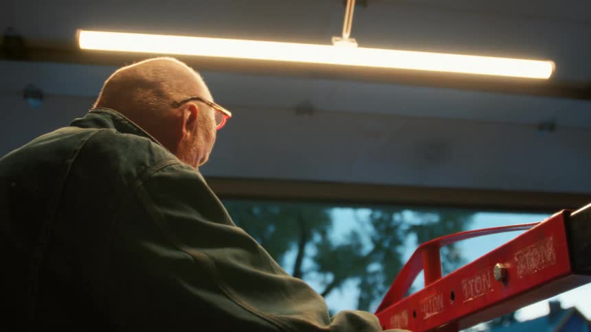 An older man with a long white beard works intently under a shop light in his home garage at dusk