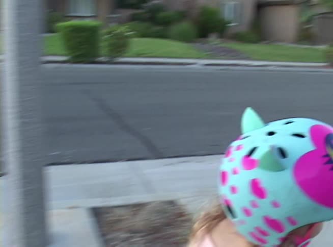 A little girl in a pink tutu and a colorful cat helmet stops for a moment before riding her scooter down the sidewalk