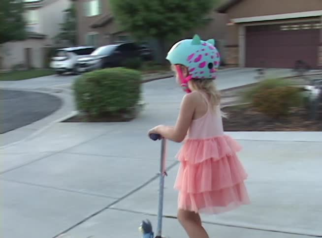 A little girl wearing a pink dress and a helmet rides her scooter down the sidewalk in a suburban neighborhood