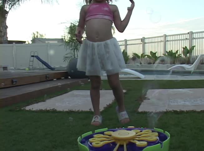 A joyful young girl with pigtails dances and plays among bubbles floating from a machine in her backyard