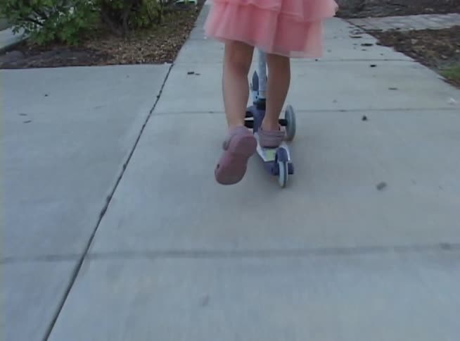 A young girl in a pink dress and a colorful cat-ear helmet rides her scooter down a suburban sidewalk